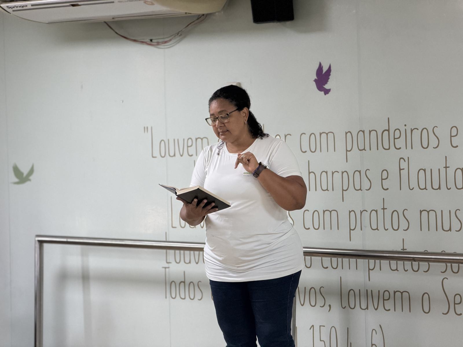 Professora conduz momento devocional, com versículo do Salmo 150 na parede